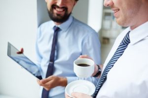 Image of young businessman holding cup of coffee while listening to his colleague at meeting